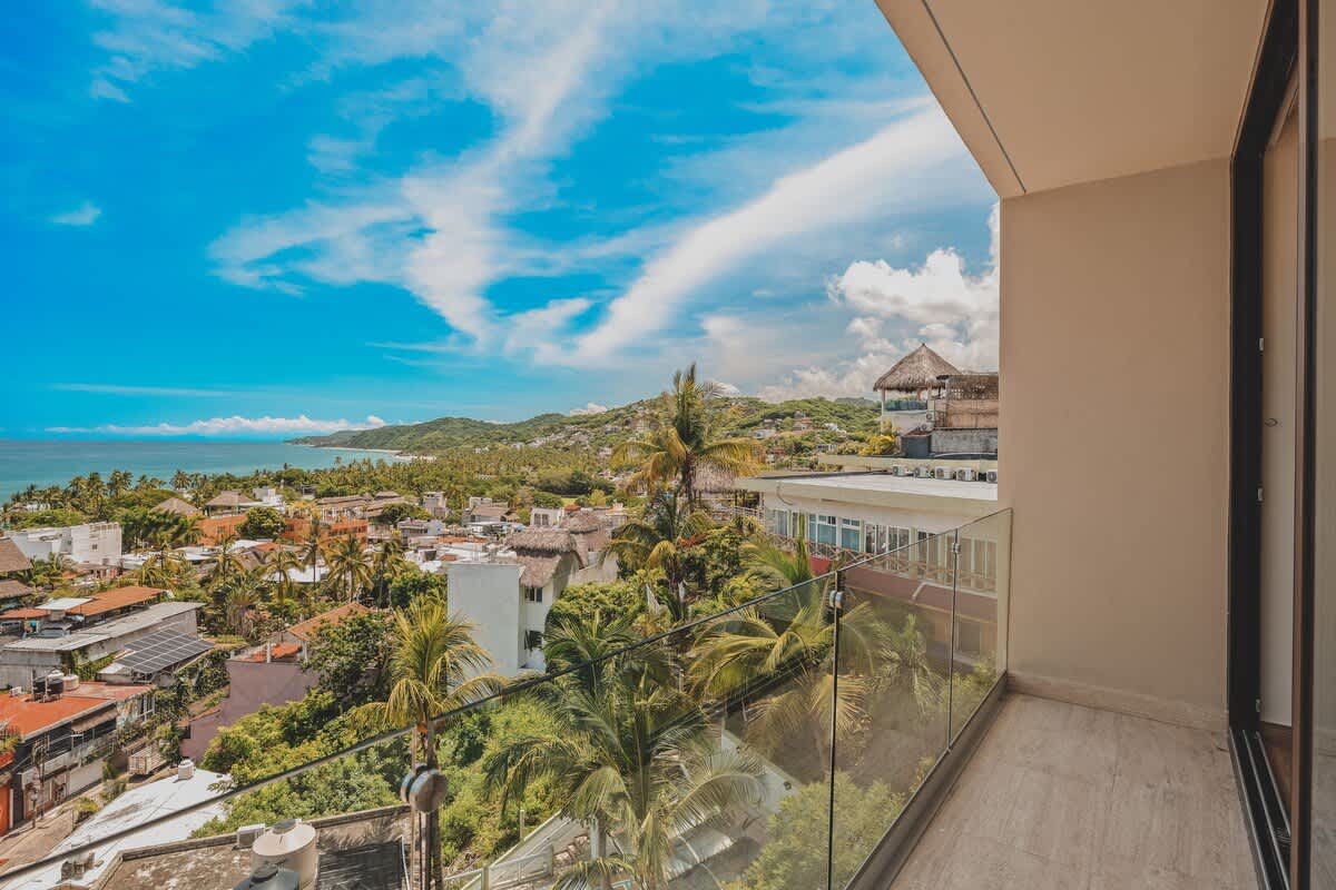 Private balcony with a glass railing, offering a stunning panoramic view of a vibrant coastal town, lush green hills, and the sparkling blue ocean under a bright sky.