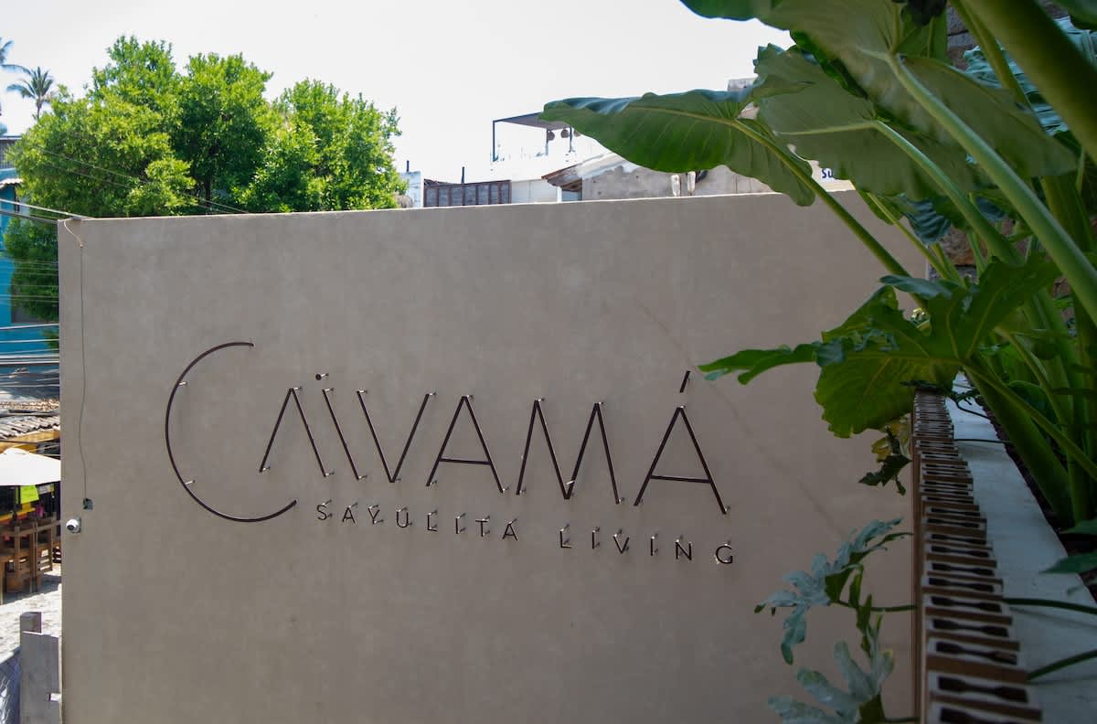 A modern metal sign spelling 'CAVAMA SAYULITA LIVING' is affixed to a light-colored wall, partially framed by vibrant green foliage, under a bright sky.