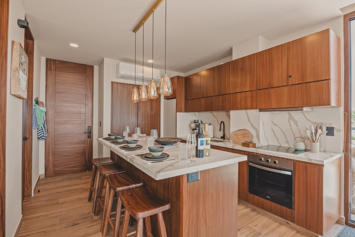 Full view of a stylish kitchen and dining area with warm wood cabinetry, white marble island and countertops, and a linear pendant light fixture, featuring a dining setup on the island and integrated kitchen appliances.