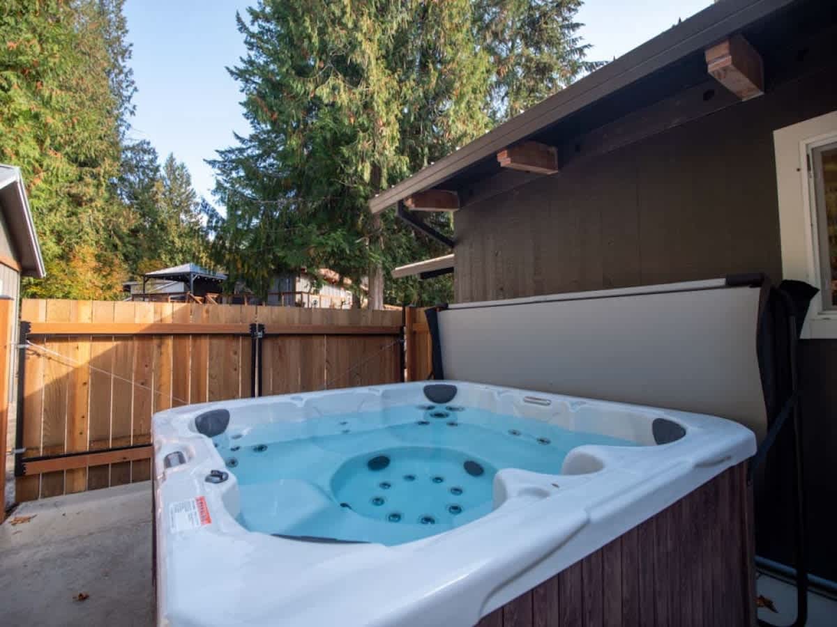 Outdoor hot tub with a light gray cover partially open, set on a concrete patio surrounded by a wooden fence and tall evergreen trees.