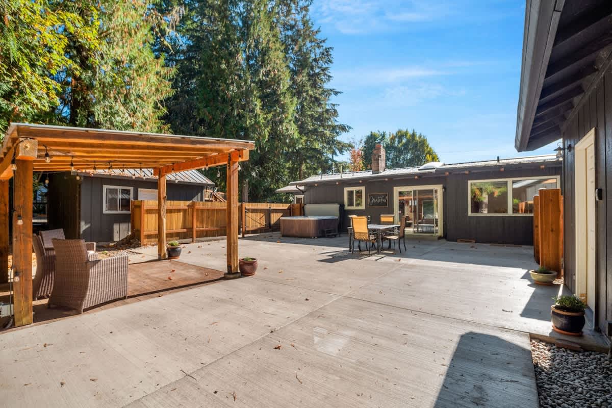 Rear exterior of a dark-colored house with large windows, overlooking a spacious concrete patio with a hot tub, outdoor dining set, and a covered pergola.