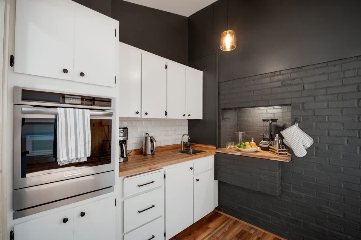 A section of the kitchen with white shaker cabinets, butcher block countertops, and a white subway tile backsplash. A stainless steel built-in oven is visible, along with a coffee maker and kettle, set against a dark grey brick wall.