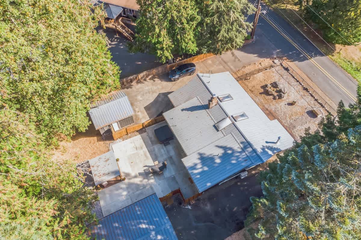 Aerial view of the rear of the house, featuring a gray metal roof, skylights, and a patio area with a dark hot tub. A small shed with a matching roof is visible nearby, all set within a wooded landscape.