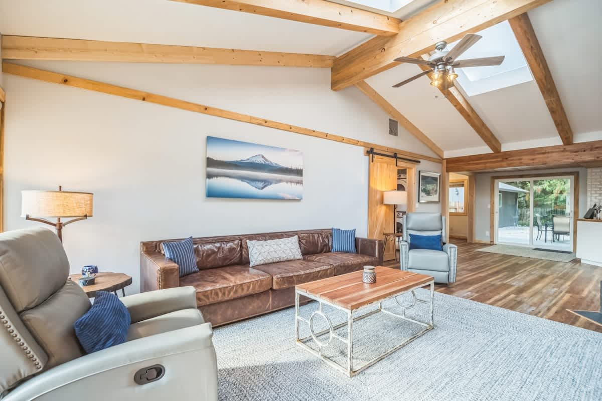 A bright living room with a vaulted ceiling, exposed wooden beams, and skylights, furnished with a brown leather sofa and two light grey recliners. A large landscape painting hangs on the wall, and hardwood floors are covered by a textured area rug.