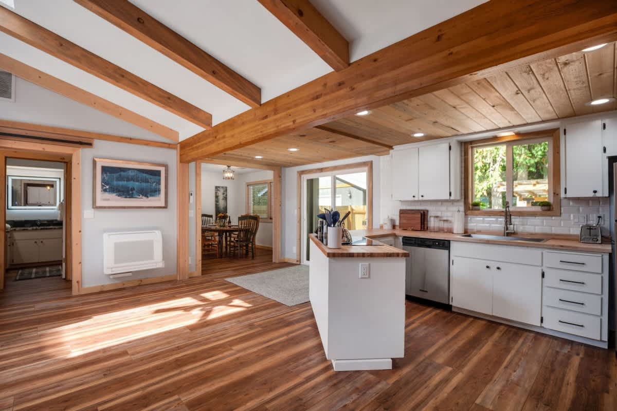 A wide shot of the kitchen and adjacent dining area, featuring white cabinets, butcher block countertops, and a natural wood paneled ceiling. Hardwood floors extend throughout, and a sliding barn door leads to another room.