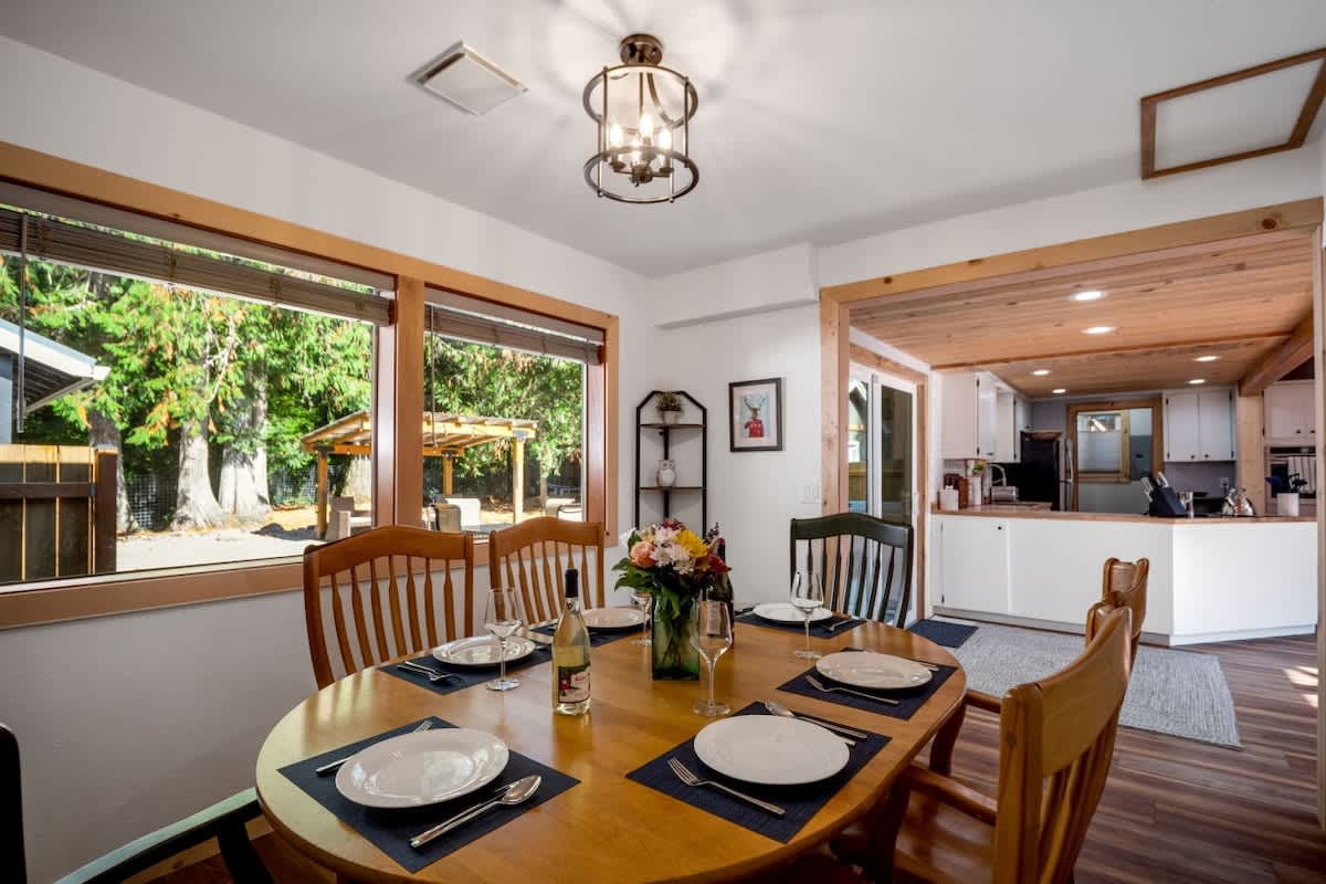 Bright dining room with an oval wooden table and mixed chairs, featuring large windows overlooking a lush green outdoor area, and an open view into the adjacent kitchen space.