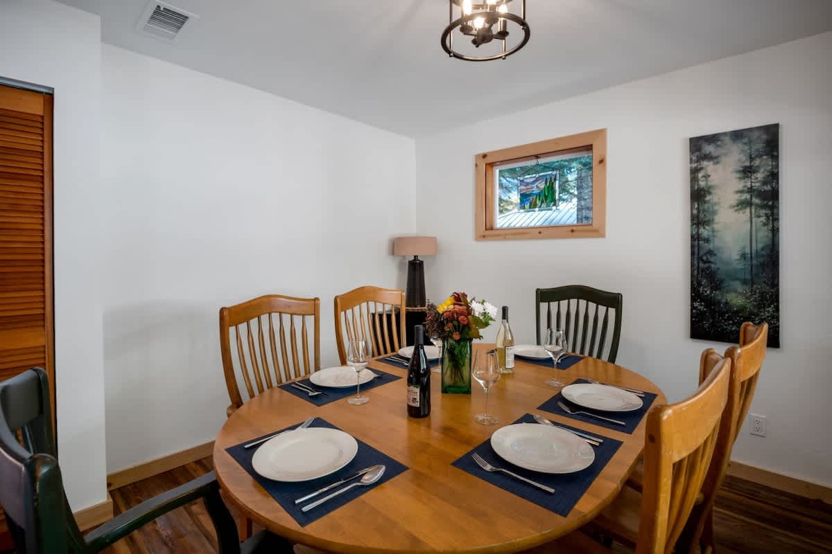 Close-up view of the dining table, set with white plates and blue placemats, featuring a floral centerpiece and wine bottles, with a glimpse of a window and a dark forest painting on the wall.