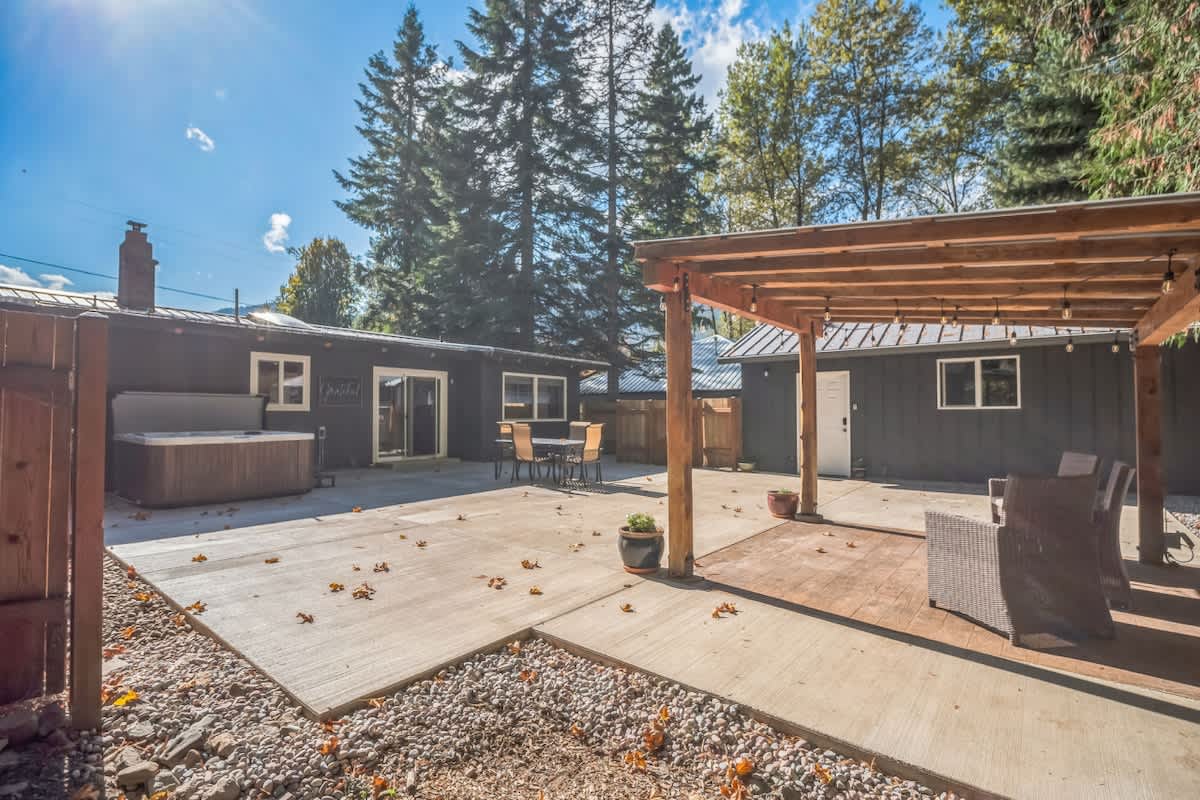 Wide view of the property's backyard, featuring a dark-colored house, a large concrete patio with a hot tub, and a wooden pergola, all set against a backdrop of tall evergreen trees.