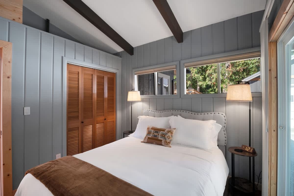 Bedroom featuring a queen bed with white bedding and a brown throw, light grey paneled walls, two nightstands with lamps, and a wooden louvered closet.