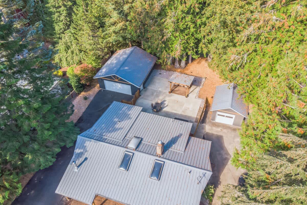 Overhead aerial view highlighting the main house with its gray metal roof and skylights, a detached garage, and a smaller shed, all with matching roofs. A wooden gazebo and paved patio area are visible between the structures, surrounded by lush evergreen trees.
