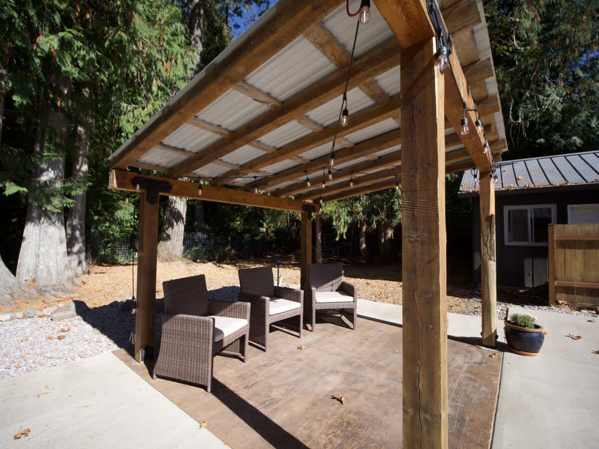 Outdoor patio area with a rustic wooden pergola covering four wicker armchairs, set on a patterned concrete slab, surrounded by tall trees.