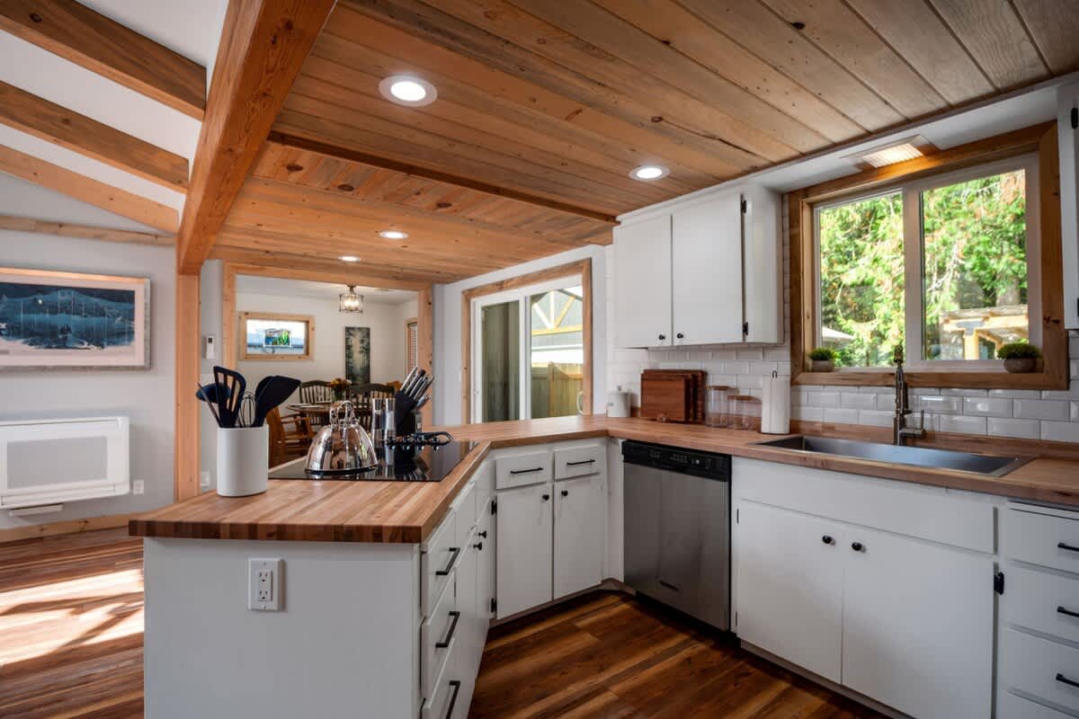 A well-lit kitchen featuring white shaker cabinets, butcher block countertops, and a white subway tile backsplash. The ceiling is paneled with natural wood, and a stainless steel dishwasher is visible next to the sink.