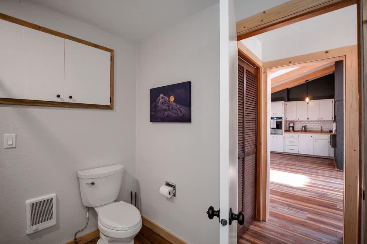 A white toilet is positioned against a light-colored wall with a small wall heater below, offering a view through a wooden doorway into a bright kitchen with white cabinets and wood floors.