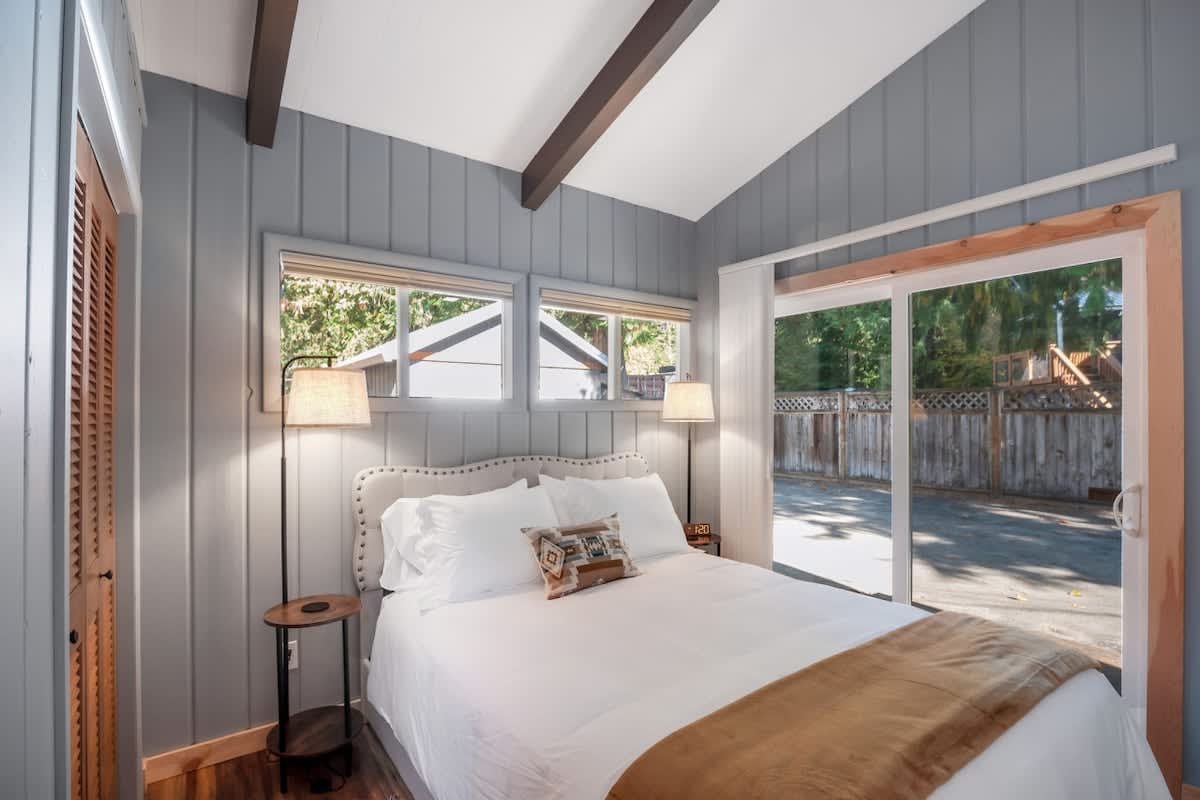 Bright bedroom with a queen bed, two modern nightstands with lamps, light grey paneled walls, and a large sliding glass door providing access to an outdoor patio area.