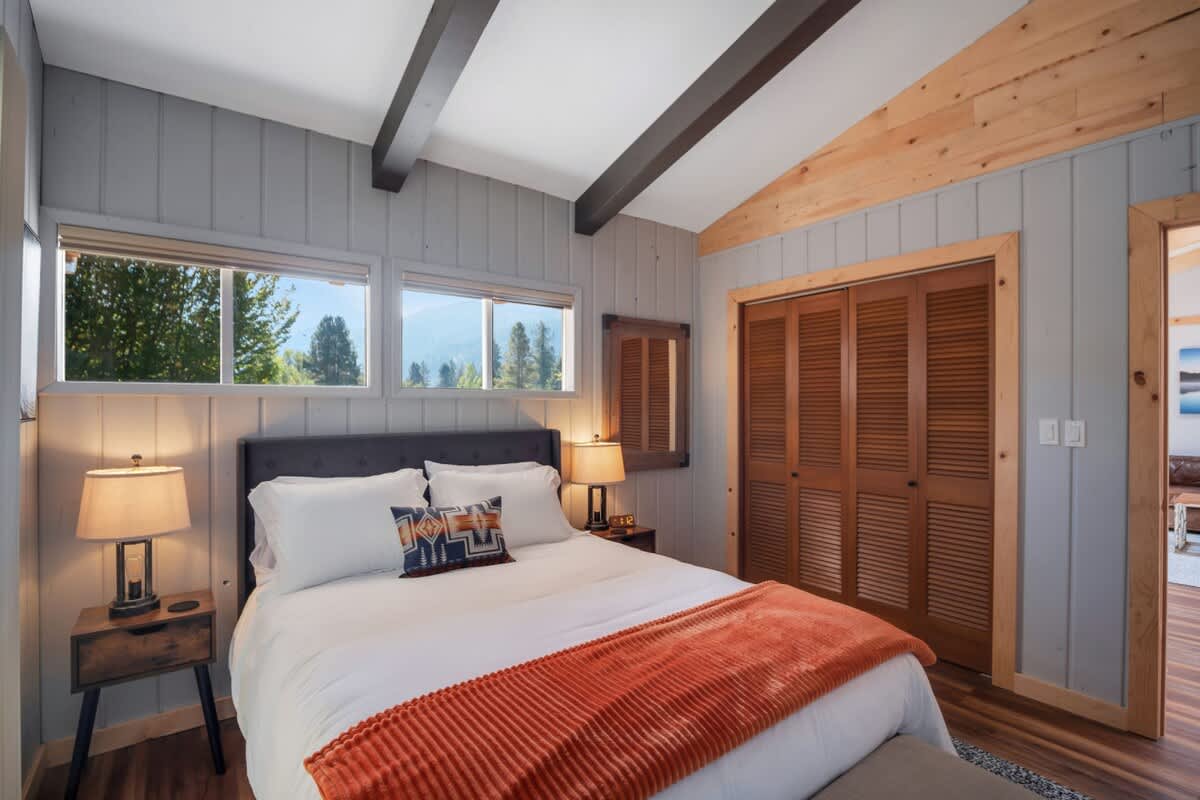 Bedroom with a queen bed, light grey paneled walls, a vaulted ceiling with dark beams, and high windows above the bed offering views of trees, alongside a wooden louvered closet and a natural wood doorway.