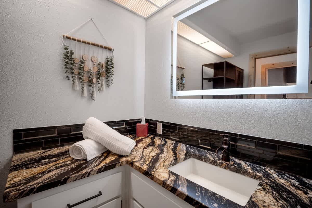 Modern bathroom vanity featuring a black and gold swirled granite countertop, a rectangular white undermount sink, and a large illuminated mirror, complemented by a decorative wall hanging with wood slices and greenery.