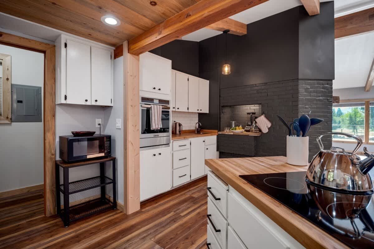 Modern kitchen with white shaker cabinets, dark countertops, and a black brick accent wall behind the stove, featuring a wooden ceiling with exposed beams and warm wood flooring.