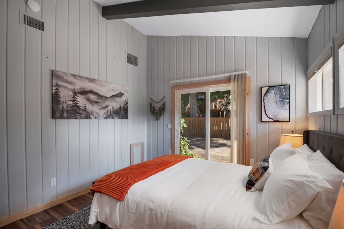 Cozy bedroom featuring a queen-sized bed with white linens and an orange throw, light grey paneled walls, a vaulted ceiling with dark exposed beams, and a sliding glass door leading to an outdoor area.