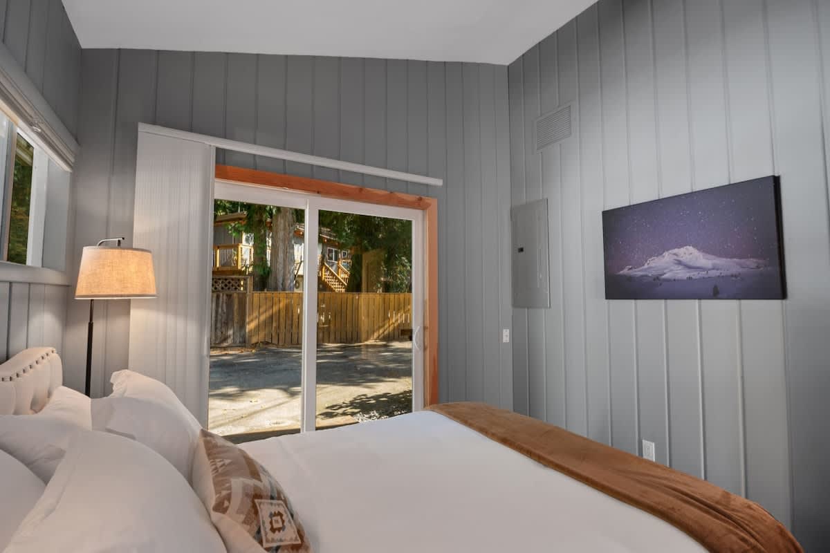 Bedroom with a queen bed, a large sliding glass door offering a view of the outdoor area, light grey paneled walls, and a wall-mounted artwork depicting a snowy mountain scene.