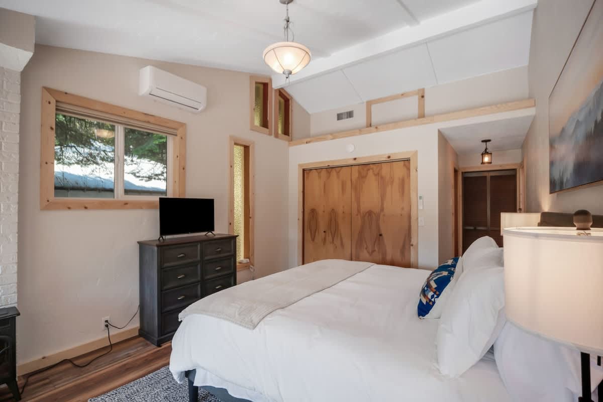 Bedroom with a queen-sized bed, dark wood dresser with a TV, and light wood-trimmed windows, including unique vertical stained-glass panels, under a vaulted ceiling with exposed beams.