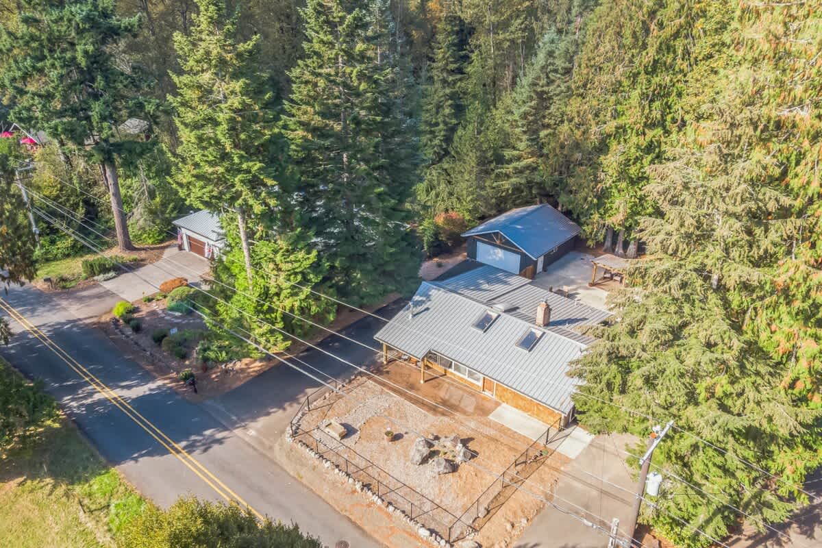 High aerial view of the property, showcasing the main house with a gray metal roof and light brown siding, surrounded by dense evergreen forests. A paved driveway leads to a detached garage, and a road runs along the front of the property.