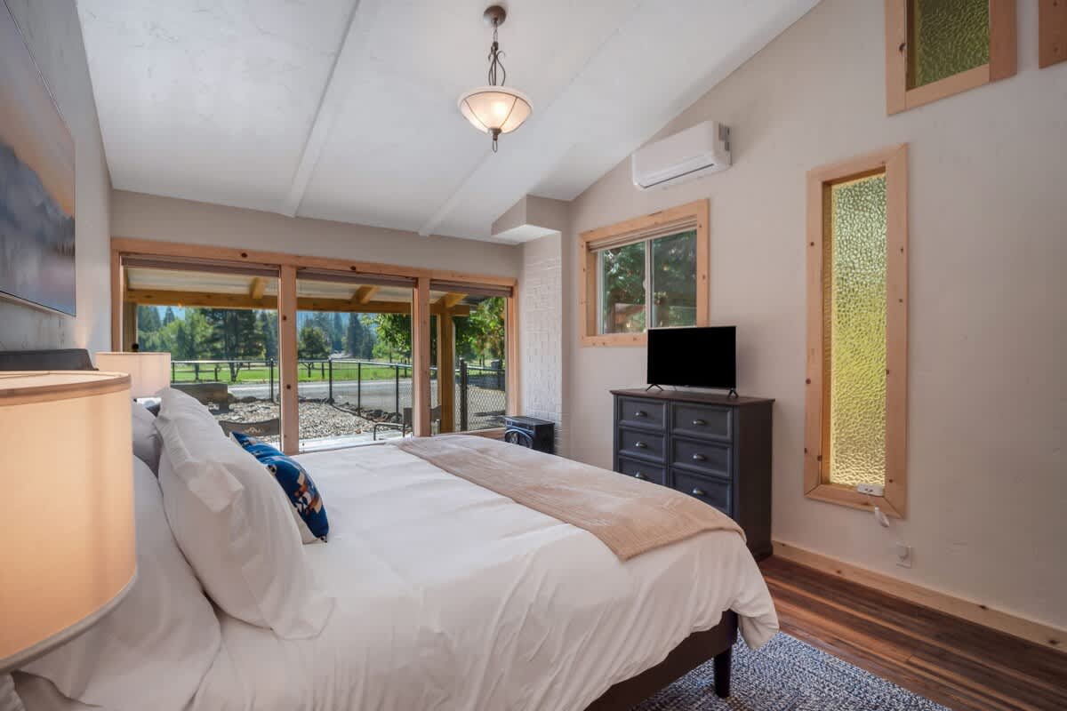 Bright bedroom with a queen bed facing large sliding glass doors that open to an outdoor view, featuring a dark dresser with a TV, and unique vertical windows with wood trim.