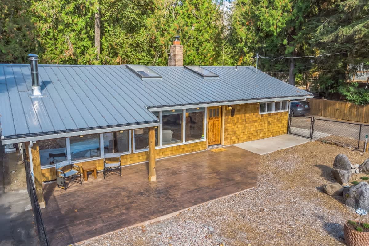 Aerial view of a single-story house with a gray metal roof, skylights, and light brown shingle siding. A large concrete patio with outdoor seating is visible in front, surrounded by gravel and tall evergreen trees.