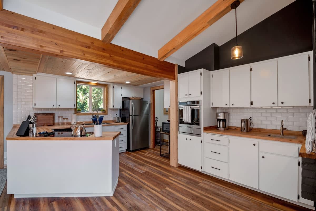 This kitchen features white shaker cabinets, butcher block countertops, and a stainless steel refrigerator and built-in oven. The space has a mix of white and dark grey walls, with hardwood floors and a natural wood beam overhead.