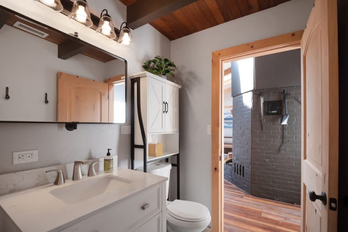 Contemporary bathroom featuring a white vanity with a rectangular sink, a large mirror, a toilet, and a white storage cabinet with barn-door style fronts, all under a wood-beamed ceiling.