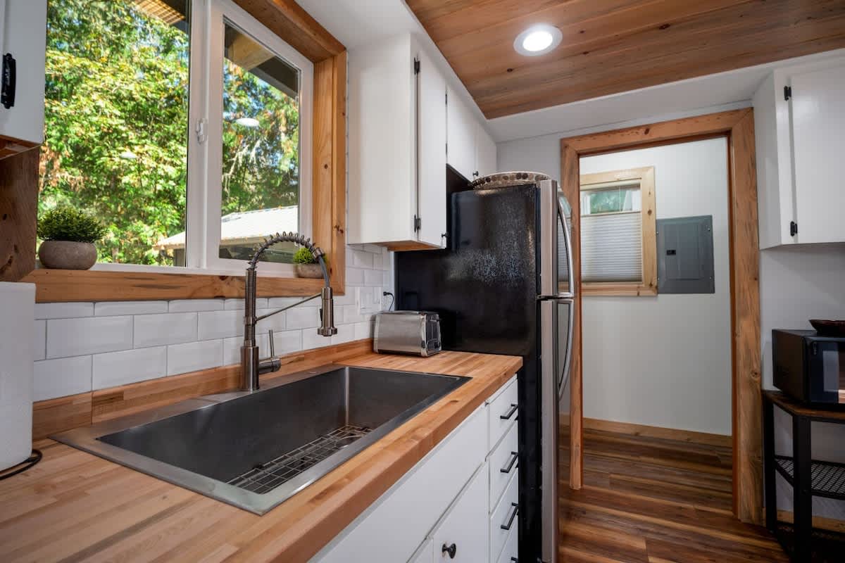 A bright kitchen corner with a stainless steel sink, butcher block countertops, and a white subway tile backsplash under a large window. A black refrigerator and toaster are visible, with natural wood trim around the window and doorway.