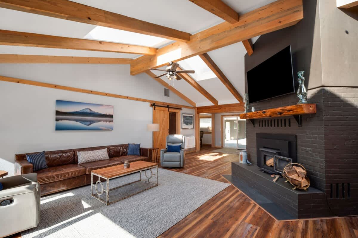 This spacious living room features a vaulted ceiling with exposed wooden beams and skylights, a large brown leather sofa, and a black brick fireplace with a mounted TV. Hardwood floors and a light-colored rug complete the cozy, rustic modern aesthetic.