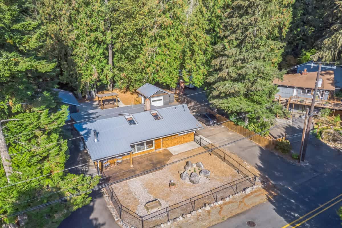Aerial view of the single-story house with a gray metal roof and light brown siding, featuring a fenced gravel area with a fire pit in the foreground. A detached garage with a matching roof is visible behind the main house, all nestled among tall evergreen trees.