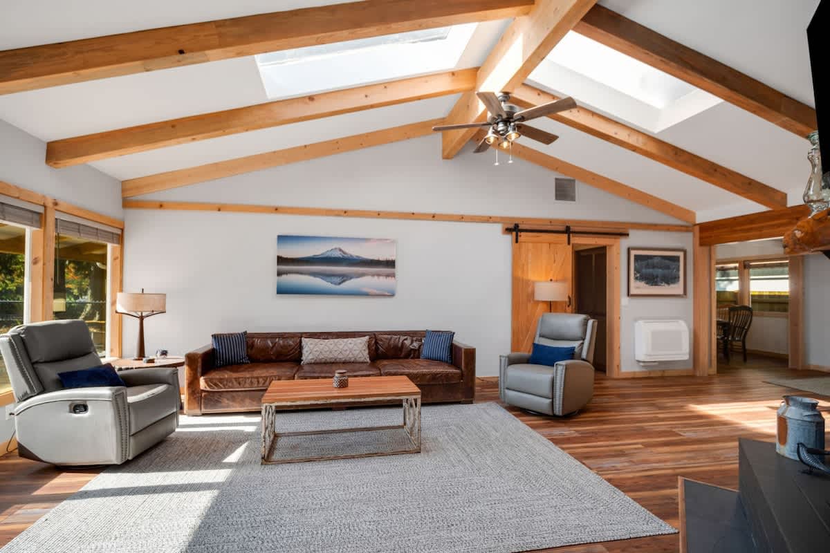 The living room features a dark brick fireplace with a wood-burning stove, a large TV mounted above, and a brown leather sofa with a coffee table on a gray rug. Vaulted ceilings with exposed beams and skylights add to the spacious feel.