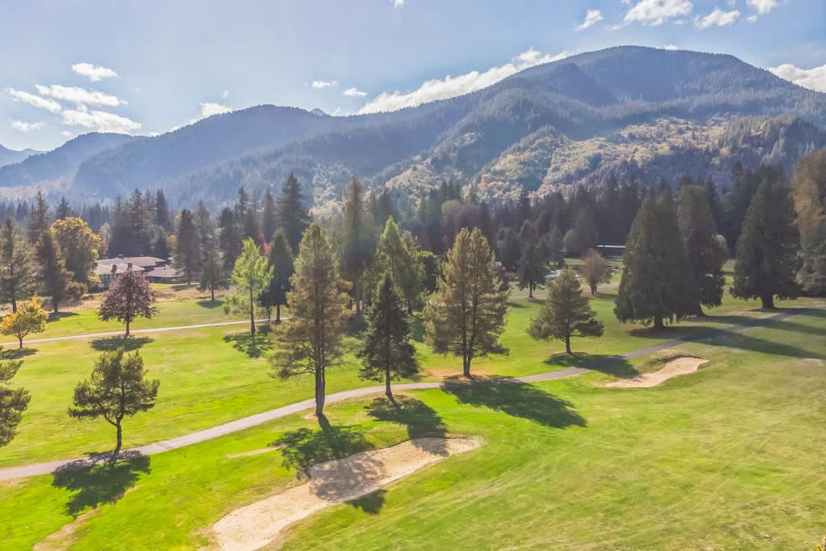 An aerial view captures a lush green golf course with sand traps and scattered trees, set against a backdrop of majestic, forested mountains under a partly cloudy sky.