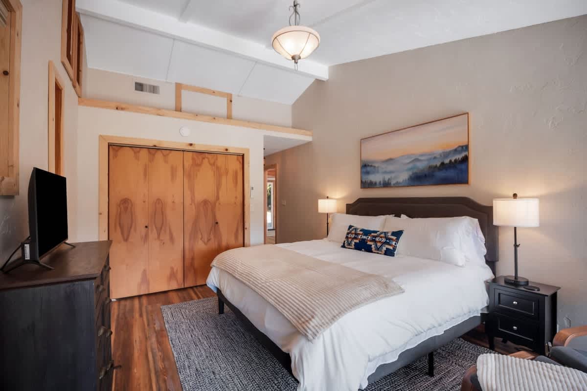 Master bedroom featuring a queen bed with white bedding and a blue accent pillow, flanked by dark nightstands with lamps, a large abstract painting above the headboard, and a wooden bi-fold closet door.