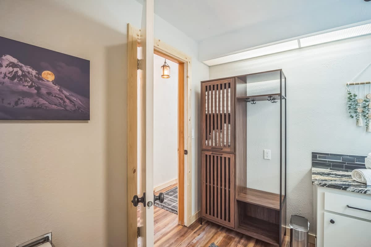 View of a bathroom with a black and gold granite vanity, a wooden slatted storage unit with hooks, and a doorway leading to another room with wood floors and a patterned rug.