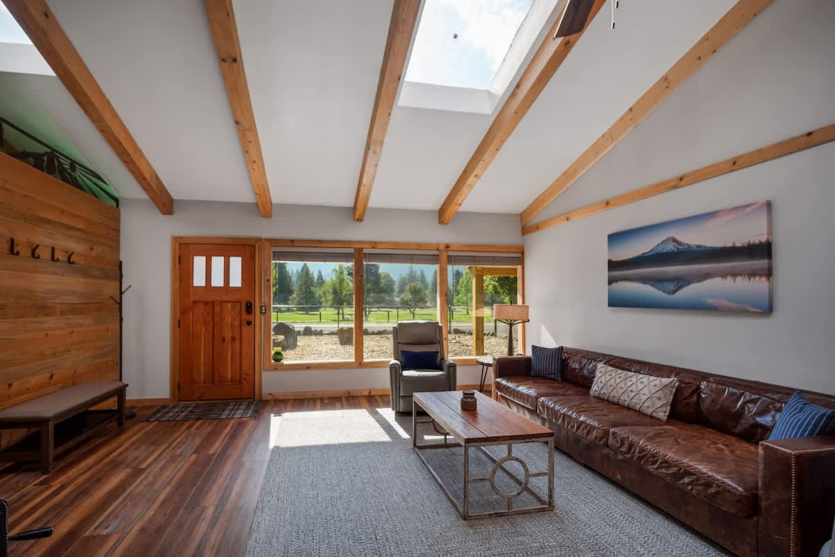 A wide shot of the living room showcasing a brown leather sofa, two gray recliners, and a coffee table on a light gray rug. The room has vaulted ceilings with exposed wooden beams and skylights, and large windows.