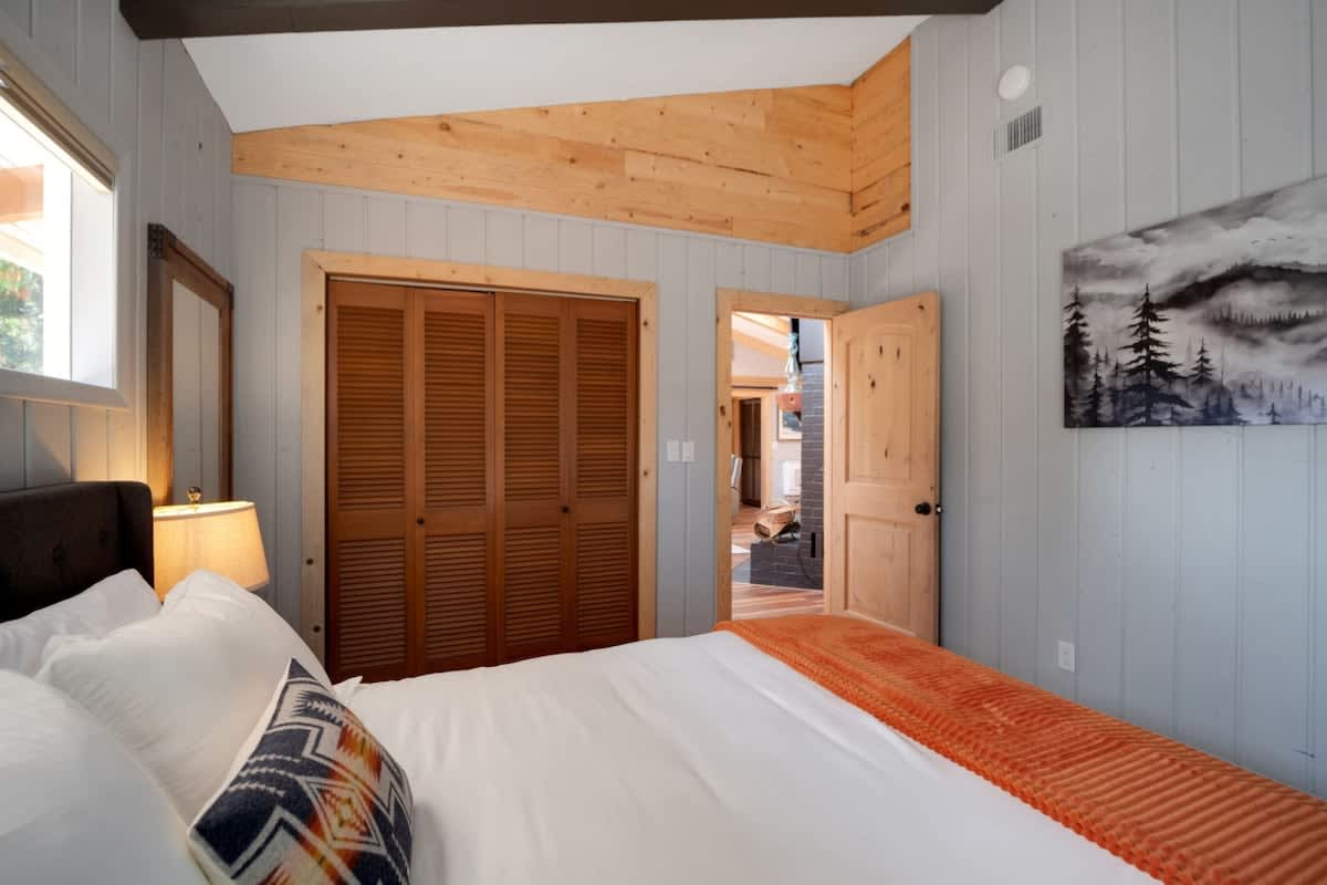 View of a bedroom with a queen bed, light grey paneled walls, a vaulted ceiling, and a prominent wooden louvered closet, with a natural wood door leading to an adjacent room.