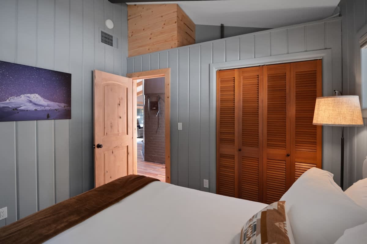 Bedroom view showing a natural wood door slightly ajar, a wooden louvered closet, and light grey paneled walls, with a mountain landscape artwork hanging above the bed.