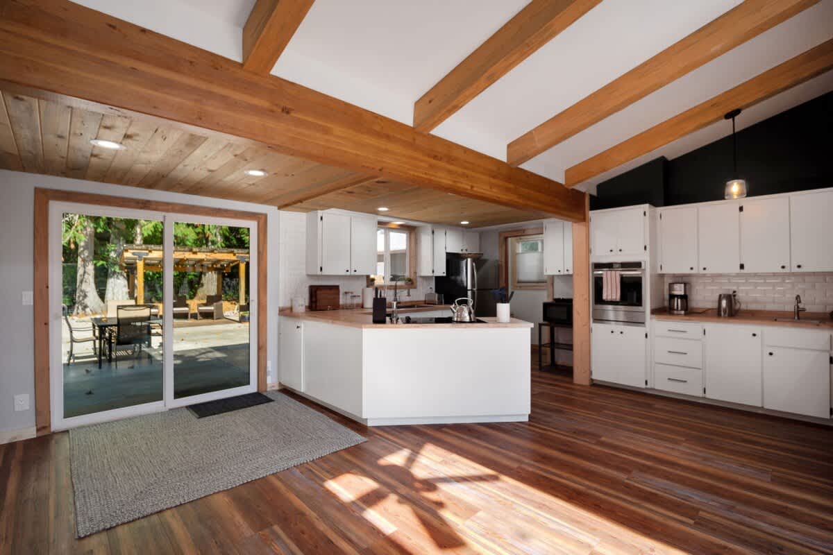 Wide view of the open-concept kitchen and dining area, showcasing white cabinetry, wood countertops, and a large sliding glass door leading to an outdoor patio, all under a vaulted ceiling with exposed wooden beams.