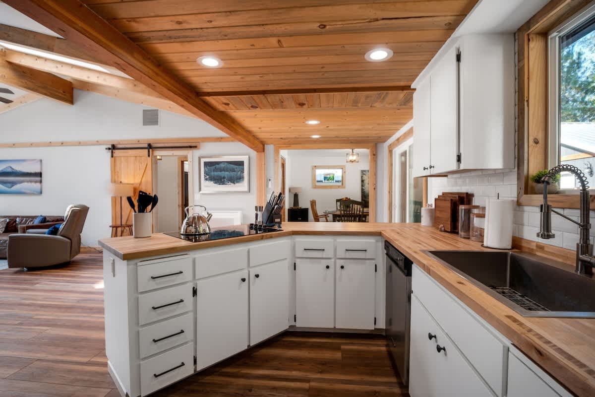 A close-up view of the kitchen island with a black induction cooktop, looking towards the living room. White shaker cabinets and butcher block countertops are prominent, with a stainless steel knife block and salt/pepper grinders on the counter.