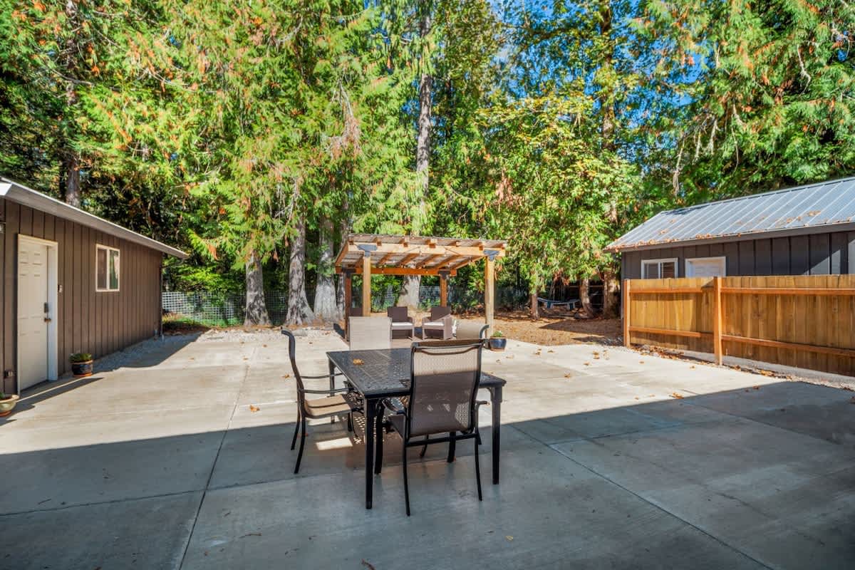 Expansive concrete patio featuring a black outdoor dining table with chairs, and a separate covered seating area under a wooden pergola, all surrounded by lush trees.
