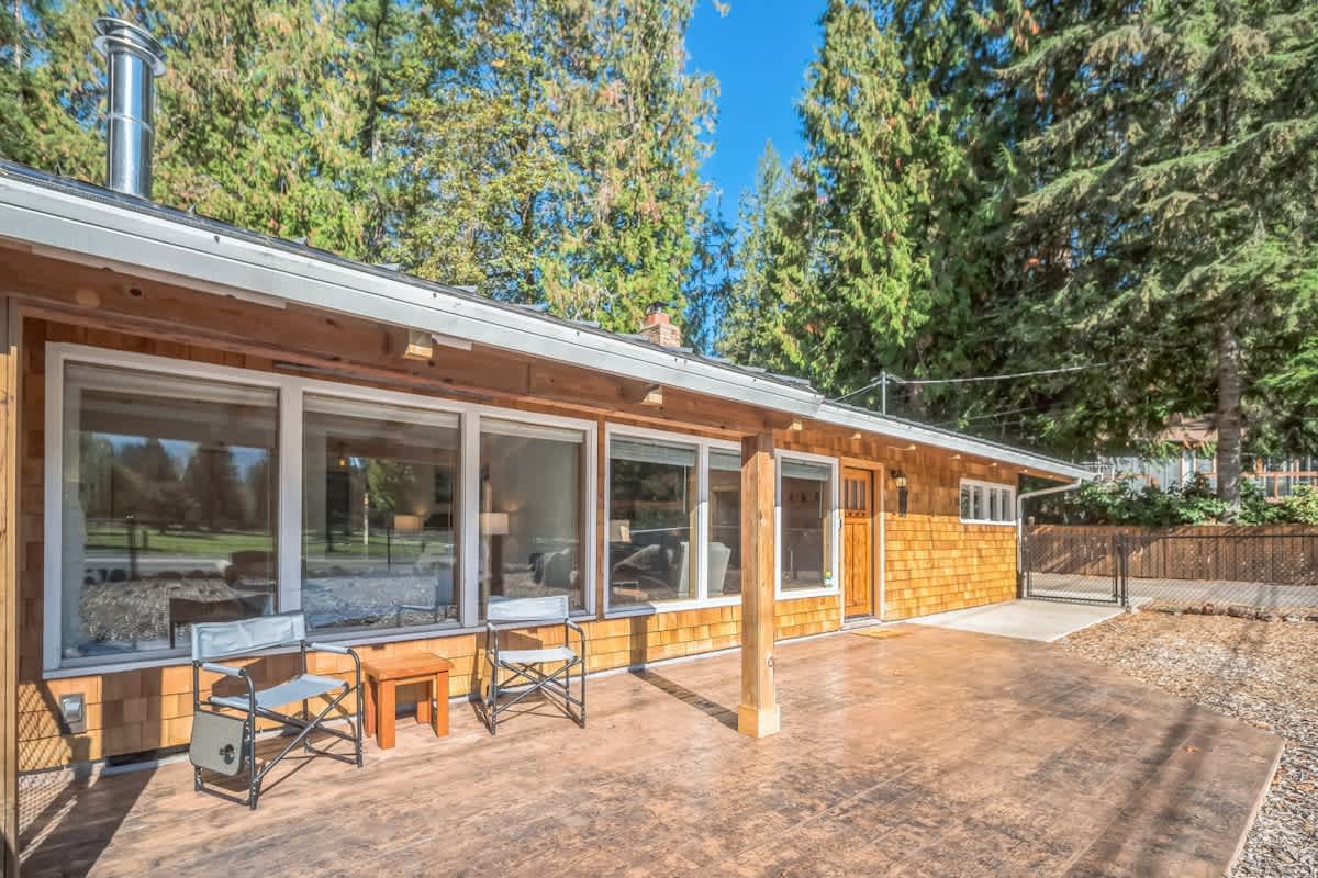 Front exterior of a house with cedar shake siding and large windows, featuring a concrete patio with two folding chairs and a small table, surrounded by tall trees.