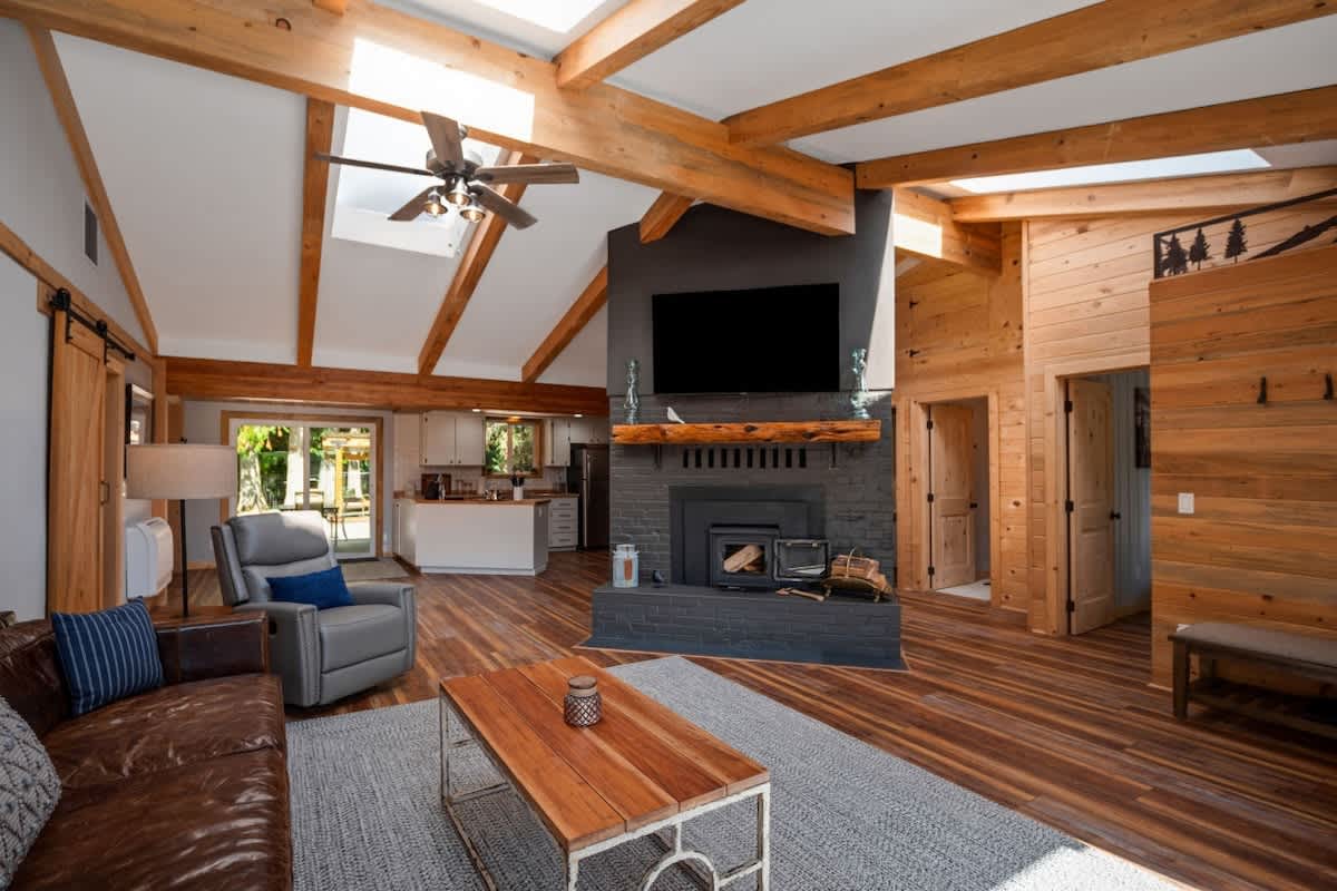 A cozy living room with a vaulted ceiling and exposed wooden beams opens to a kitchen area. A prominent dark brick fireplace with a wood-burning stove and a mounted TV serves as a focal point. Hardwood floors run throughout.