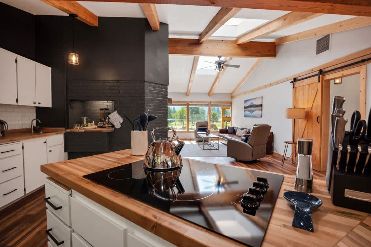 This image captures a modern kitchen island with a black induction cooktop, looking towards a black brick fireplace and the living room beyond. White shaker cabinets and butcher block countertops contrast with the dark fireplace.