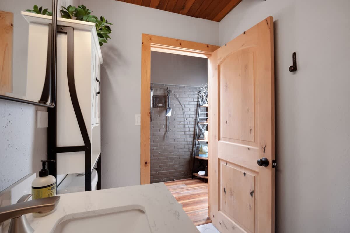 View of the bathroom vanity and toilet, with a natural wood door open to reveal an adjacent room with a dark brick wall and wood flooring.