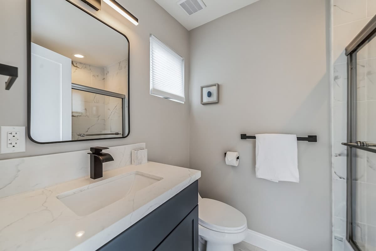 Modern bathroom with a dark blue vanity, white marble-patterned countertop, rectangular sink, and black fixtures, featuring a large rectangular mirror and a glass-enclosed shower.