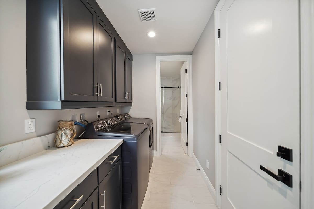 A narrow laundry room features dark gray cabinets, white quartz countertops, and a front-loading washer and dryer set. White tiled flooring contrasts with the light gray walls, and a doorway leads to an adjacent bathroom.
