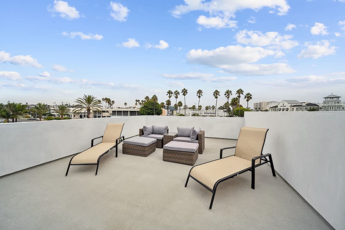 Ground-level outdoor deck area with two beige lounge chairs and a separate seating arrangement featuring a small wicker sofa and ottomans, set against a white wall with distant palm trees.
