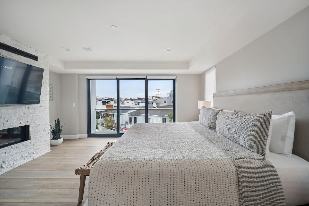 Modern bedroom with a large bed, light wood flooring, and a white stacked stone fireplace with a mounted TV above it, opposite large sliding glass doors.